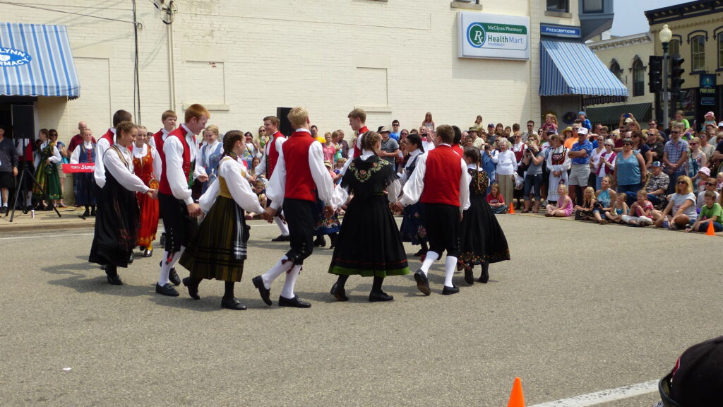 A group of people in traditional clothing perform a folk dance in a circle on a street during Syttende Mai, watched by a large crowd. , The Goose Crown Inn