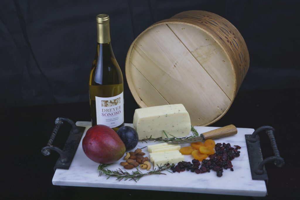 A marble platter with cheese, dried apricots, cranberries, almonds, a mango, a plum, rosemary sprigs, a wine bottle, and a round wooden box on a black background. , The Goose Crown Inn