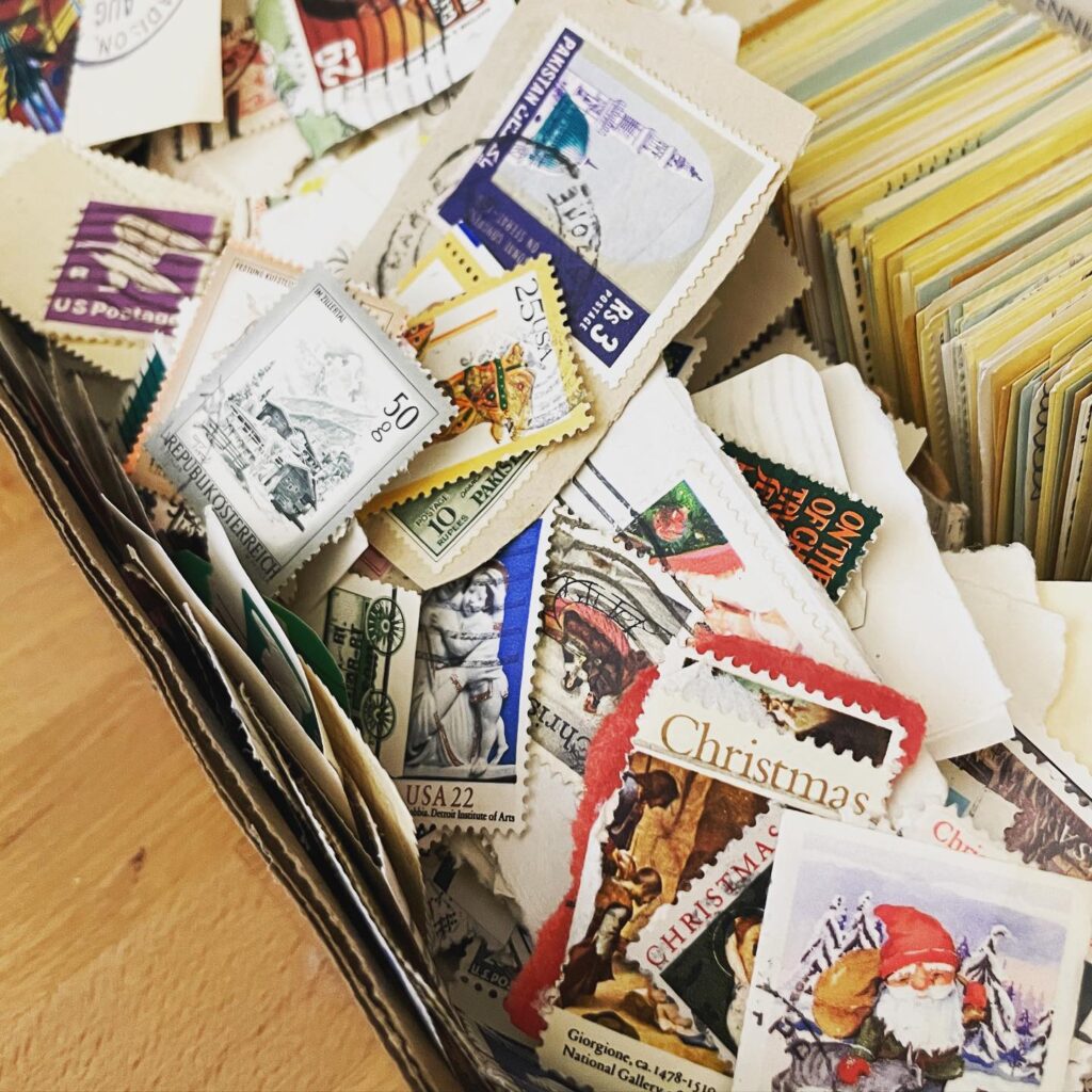 A close-up of a box filled with assorted postage stamps, including holiday-themed and vintage designs, some stacked and others scattered. , The Goose Crown Inn