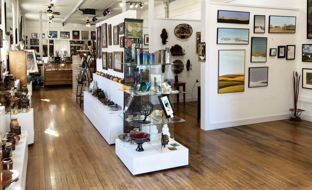 Art gallery interior with paintings on white walls, glass display shelves with ceramics and glassware, and wooden floors under bright ceiling lights. , The Goose Crown Inn