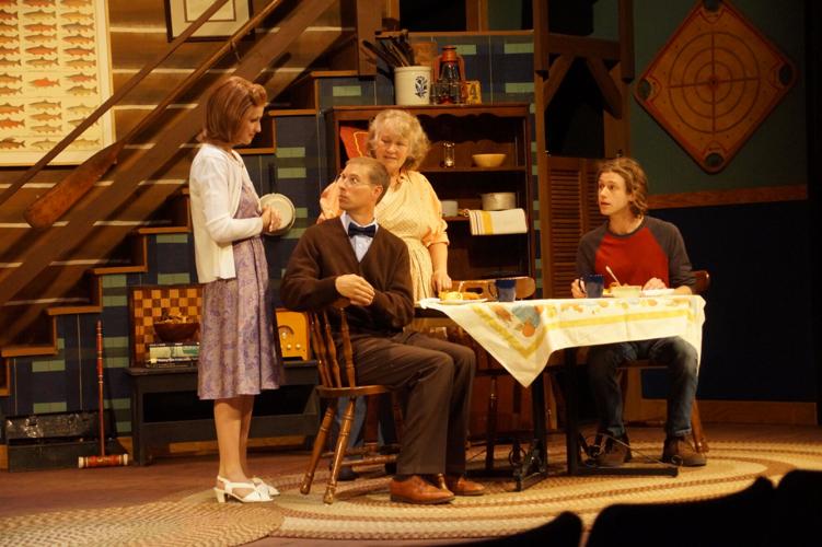 Four people sit and stand around a table set for a meal in a cozy, decorated room, suggesting a family gathering or conversation in a theatrical stage setting. , The Goose Crown Inn