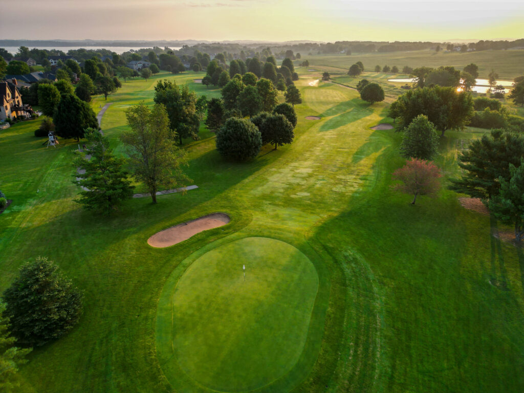Aerial view of a golf course at sunrise, showing a green, sand bunkers, fairways, trees, and distant water. , The Goose Crown Inn