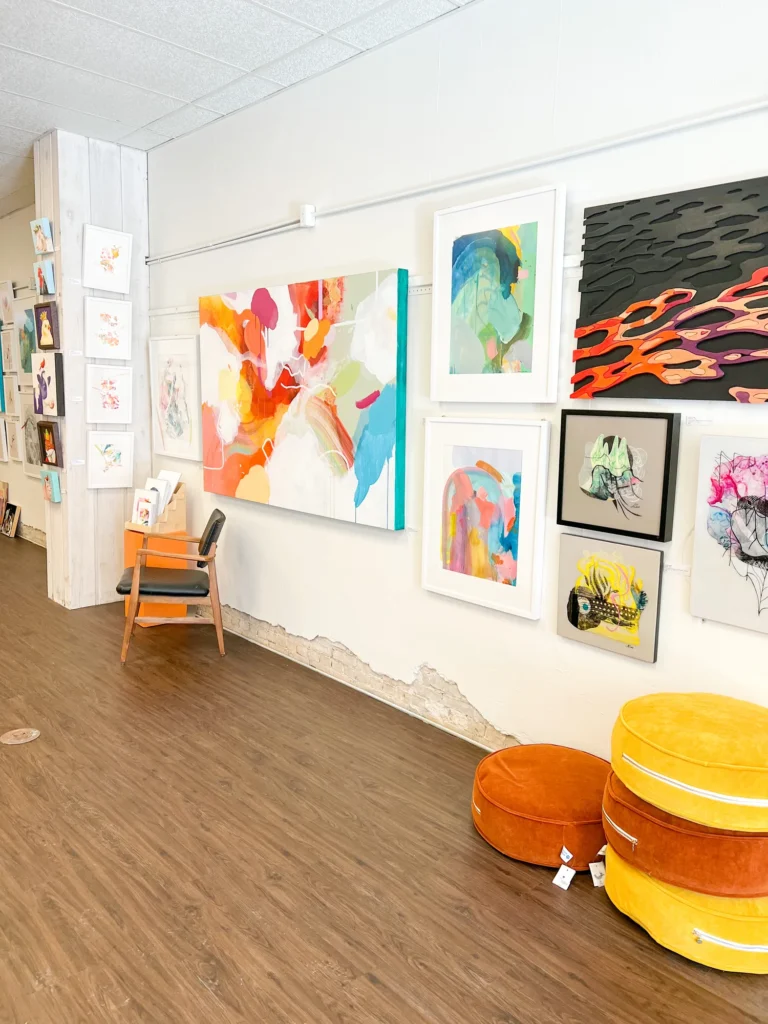 A white gallery wall displays colorful abstract and portrait artworks above wood flooring, with a chair and two orange round cushions in the foreground. , The Goose Crown Inn