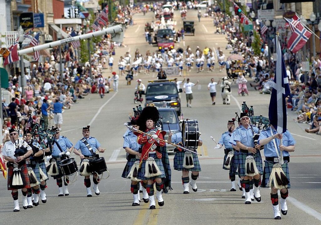 A marching band in kilts and uniforms leads a parade down a crowded street lined with spectators and flags. , The Goose Crown Inn