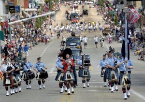 A marching band in kilts and uniforms leads a parade down a crowded street lined with spectators and flags. , The Goose Crown Inn