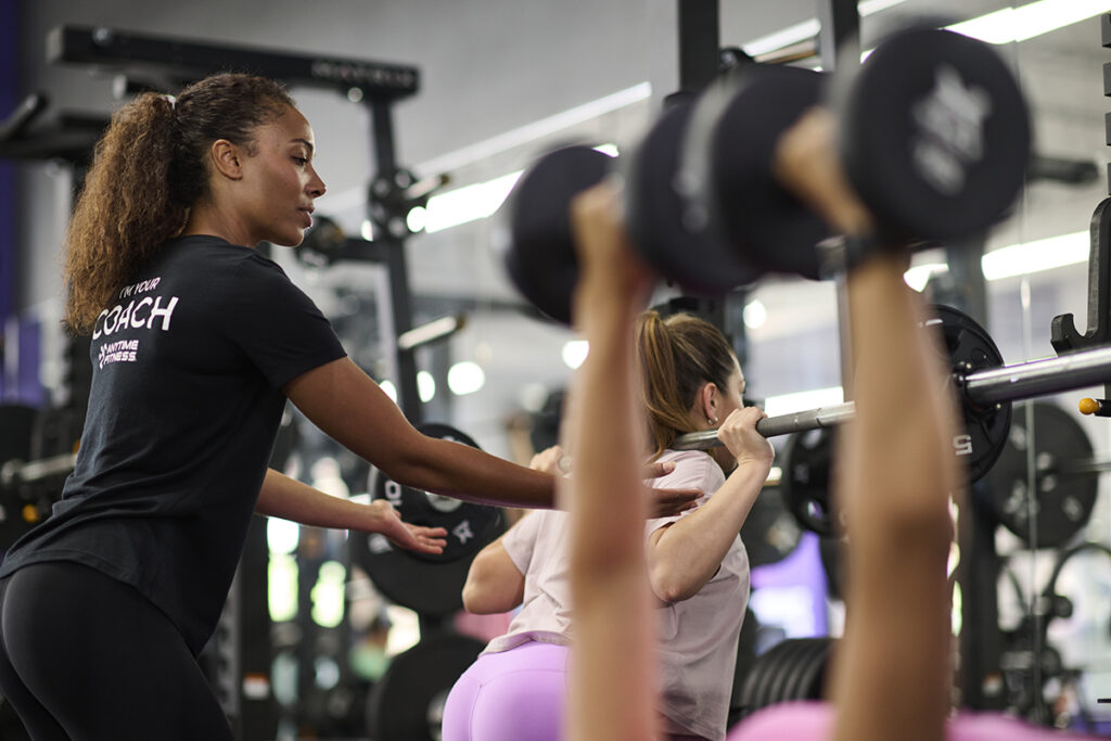 A fitness coach guides a woman performing a barbell squat in a gym, while another person lifts dumbbells in the foreground. , The Goose Crown Inn