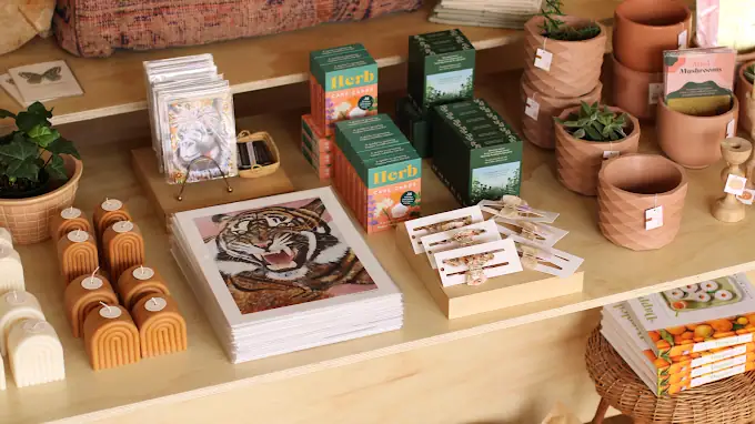 Display shelf with tiger art prints, “Herb” growing kits, potted plants, candles, greeting cards, small gift boxes, and magazines neatly arranged. , The Goose Crown Inn