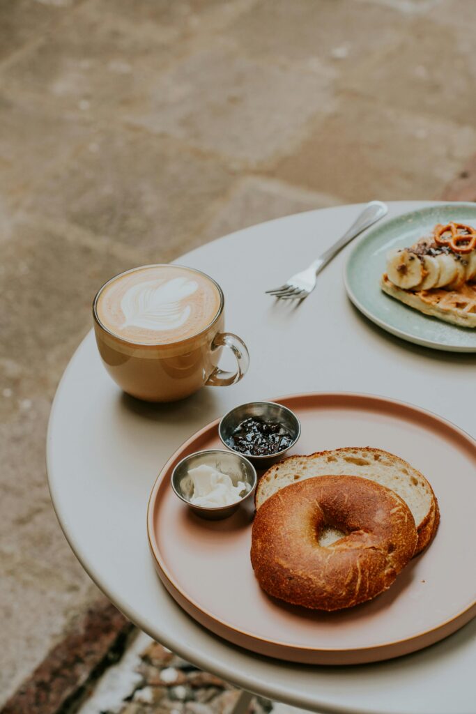 A bagel with cream cheese and jam, and a cup of latte on a round table; another plate with sliced fruit and pastry is partially visible. , The Goose Crown Inn