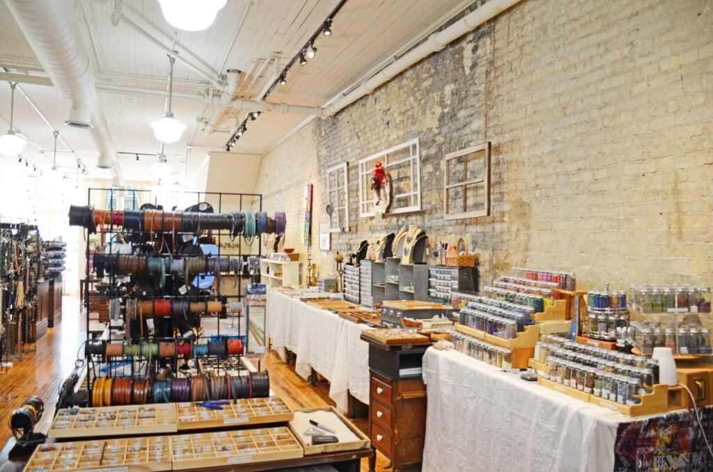 Interior of a bead and jewelry supply store with display tables, shelves of beads, rolls of leather cords, and jewelry-making tools arranged neatly. , The Goose Crown Inn