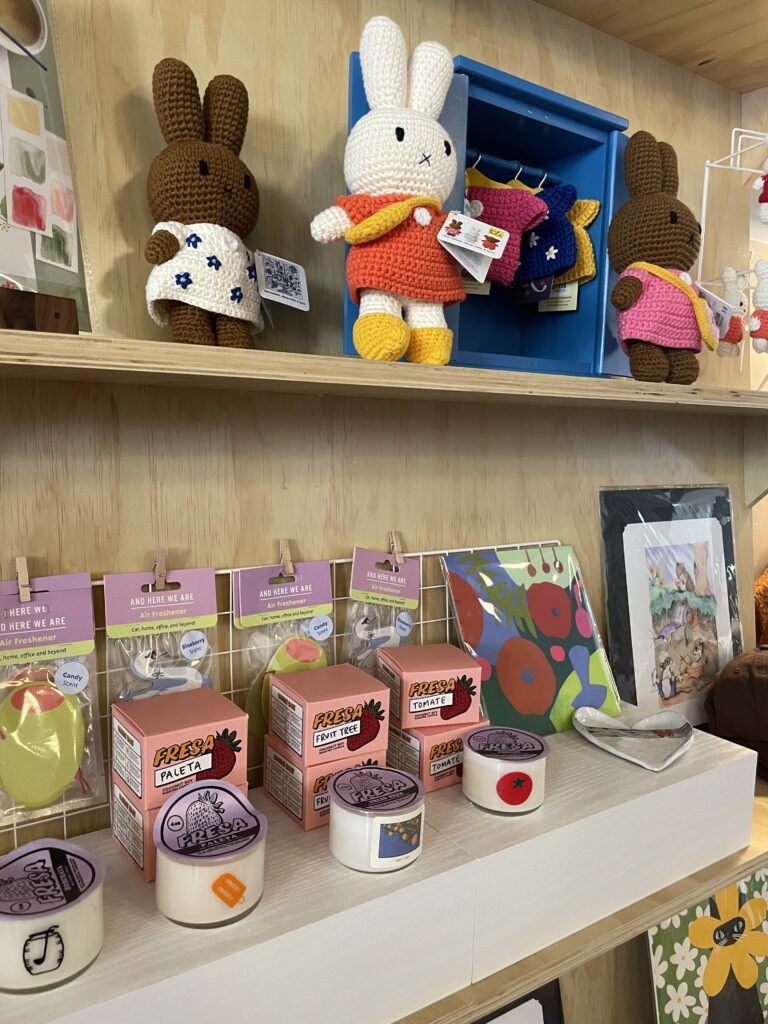 A display shelf with crocheted bunny dolls, decorative tapes, small boxes, and various colorful art prints and items arranged neatly. , The Goose Crown Inn