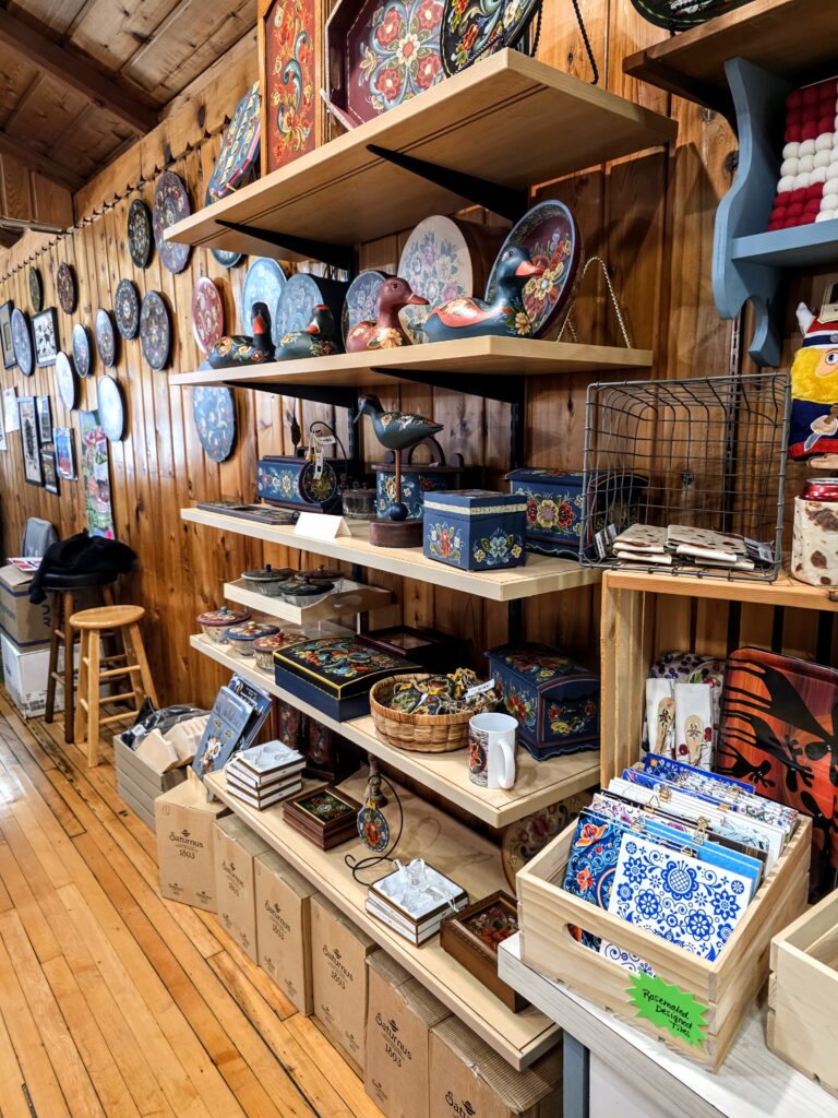 Shelves display colorful, traditional folk art items, including painted plates, wooden boxes, trays, and textiles inside a wooden-paneled gift shop. , The Goose Crown Inn
