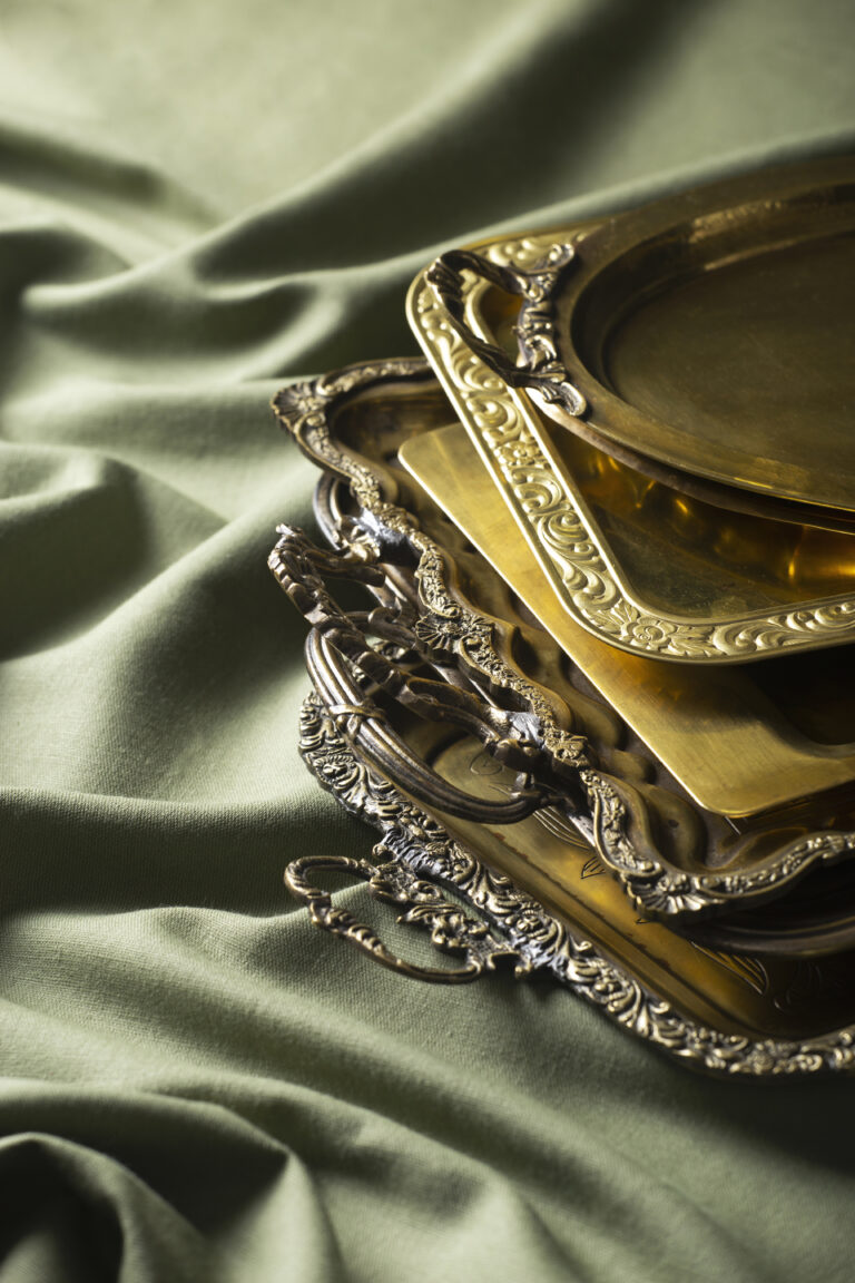 A stack of ornate gold serving trays with decorative edges, placed on a green fabric background. , The Goose Crown Inn
