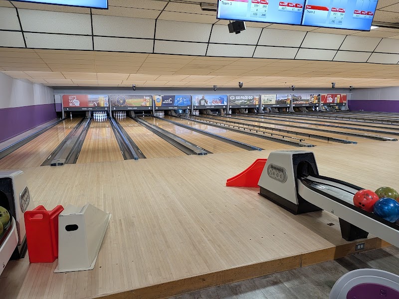 A bowling alley with several empty lanes, a bowling ball return rack holding colorful balls, and a red ramp for assisted rolling. Scoreboards are visible above the lanes. , The Goose Crown Inn