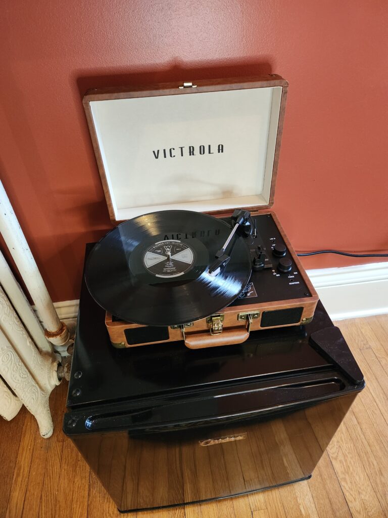 A Victrola record player with an open lid plays a vinyl record, sitting on top of a black mini fridge against a red wall. , The Goose Crown Inn