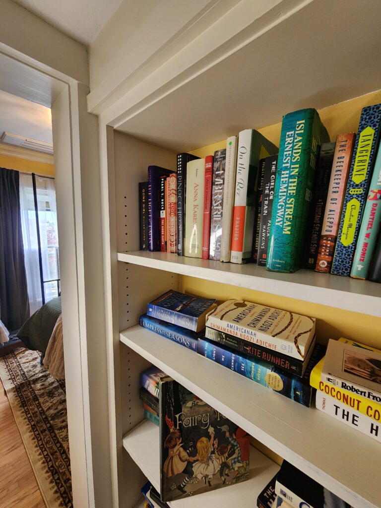 A bookshelf with three shelves holds various books, including "Island in the Stream," "The Kite Runner," and a children's book titled "Ferry" at the bottom. A bedroom is visible through a doorway. , The Goose Crown Inn