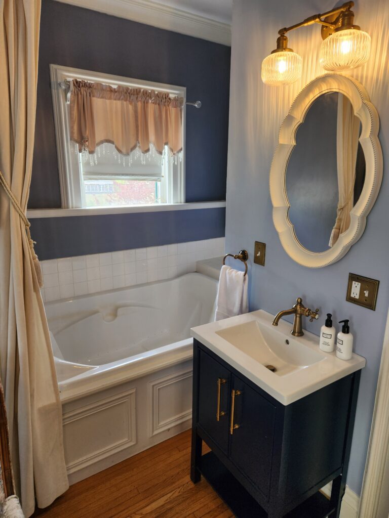 Bathroom with a white spa tub, a window with a pink valance, a navy vanity with a sink, gold fixtures, a mirror, and two glass light fixtures on a blue wall. , The Goose Crown Inn