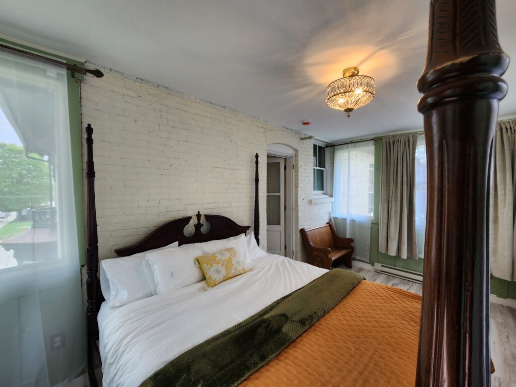 A bedroom with a four-poster bed, white brick wall, gold light fixture, curtained windows, an armchair, and a green blanket over an orange quilt. , The Goose Crown Inn