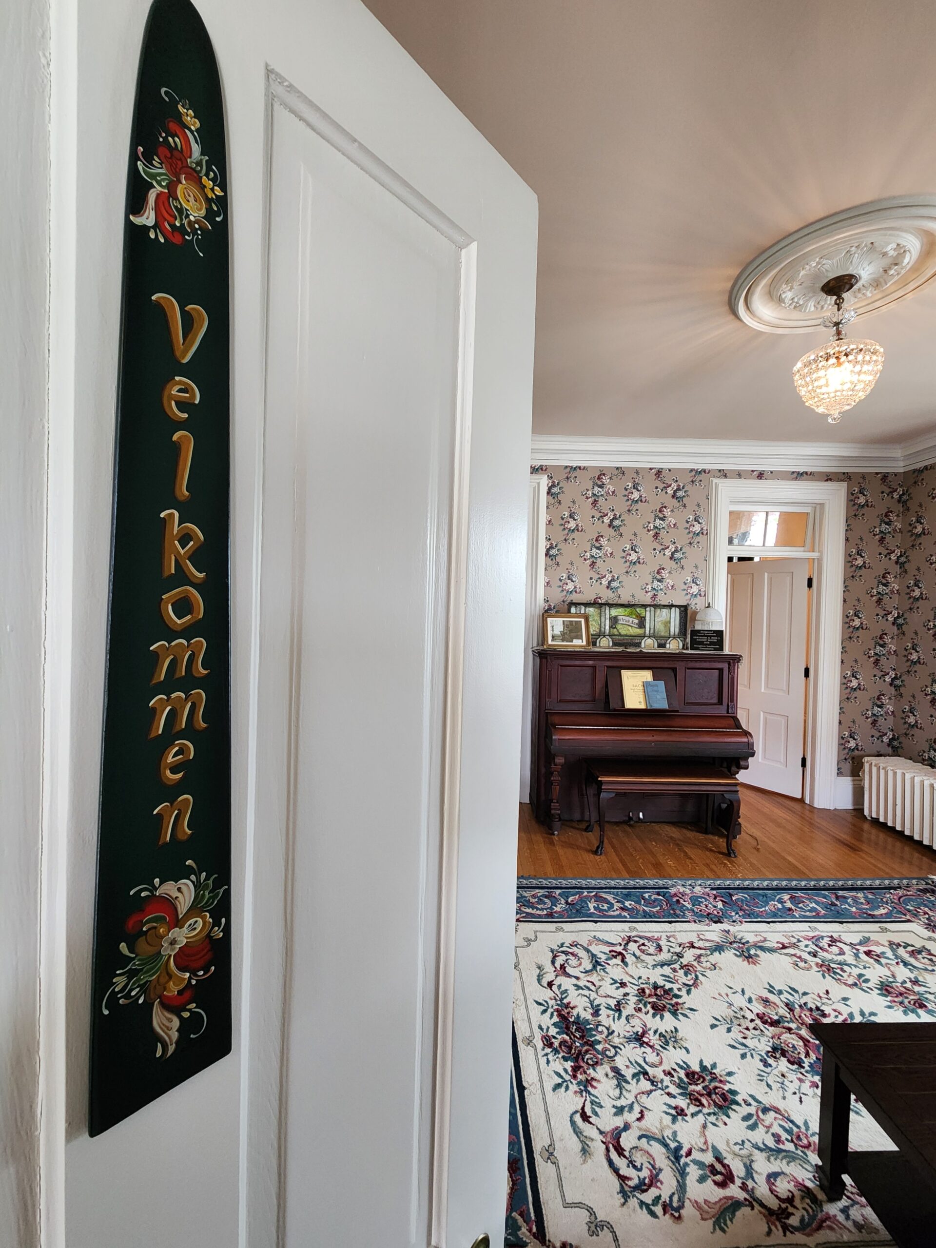 A white door partially open reveals a room with floral wallpaper, a piano, a chandelier, and a rug; a wooden sign on the door reads "Velkommen. , The Goose Crown Inn