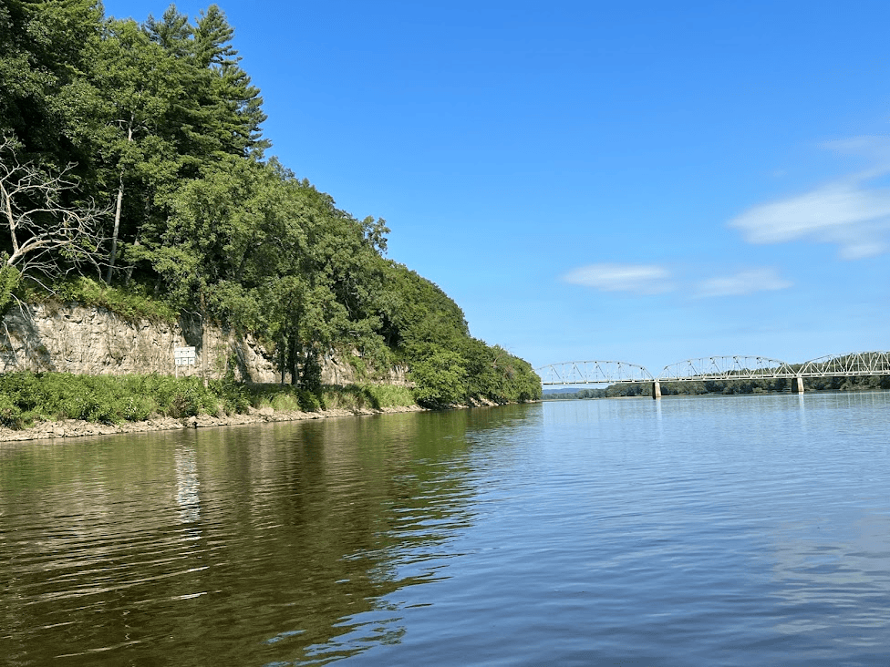 A calm river with a tree-lined rocky shoreline on the left and a metal bridge spanning the water in the distance under a clear blue sky. , Southern Wisconson Activities