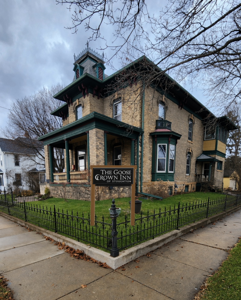A two-story brick Victorian-style house with green trim, a porch, and a sign reading "The Goose Crown Inn of Watertown," surrounded by a black metal fence on a cloudy day. , The Goose Crown Inn