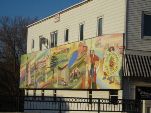 Colorful mural on building wall depicts rural scenes and people in traditional Scandinavian attire, with the text "Velkommen Stoughton. , The Goose Crown Inn