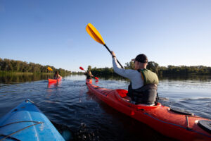Three people kayaking on a calm river, wearing life vests and paddling in the same direction on a clear day with blue sky. , Kayaking the Yahara River