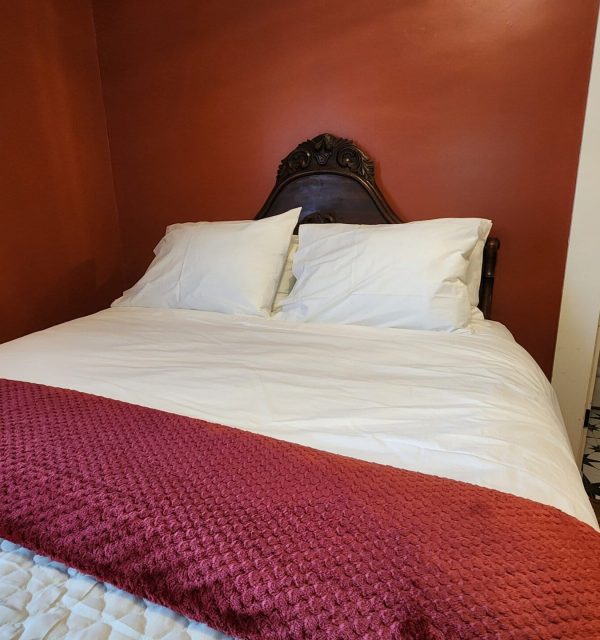 A made bed with white sheets and pillows, a dark wooden headboard, and a red textured blanket, next to a bathroom with a patterned tile floor. , The Goose Crown Inn