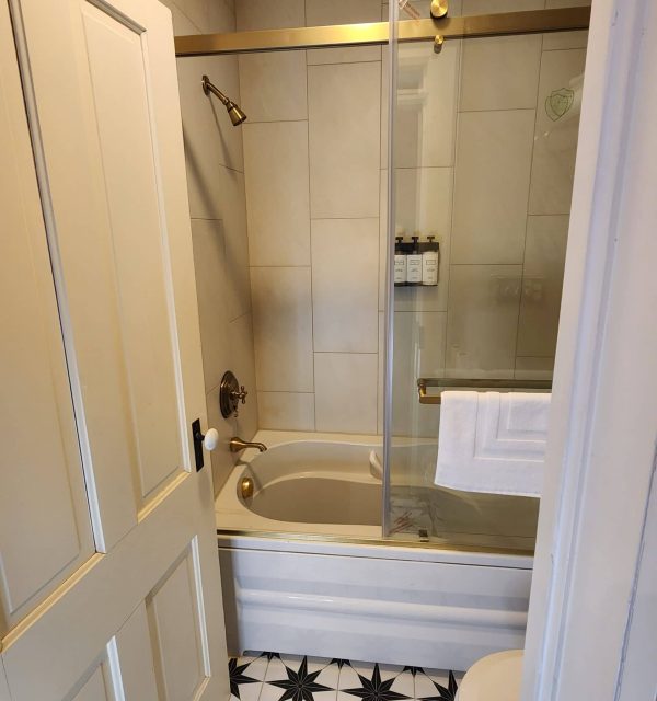 Small bathroom with a white bathtub, glass shower door, gold fixtures, wall-mounted toiletries, white tile walls, black and white patterned floor, and a partially open door. , The Goose Crown Inn