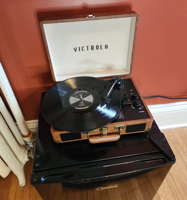 A Victrola record player with an open lid plays a vinyl record, sitting on top of a black mini fridge against a red wall. , The Goose Crown Inn