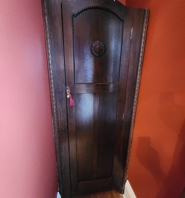 A tall, dark wooden corner cabinet with ornate detailing, set against red walls and a hardwood floor, with a small pink tassel hanging from the keyhole. , The Goose Crown Inn