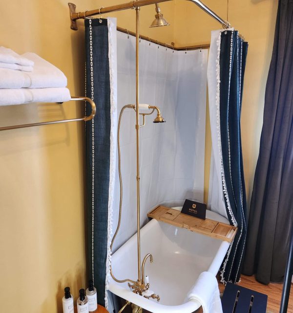 A vintage-style bathroom with a clawfoot tub, overhead shower, shower curtain, towel rack with folded towels, a wooden stool with bottled toiletries, and a small wooden bath tray. , The Goose Crown Inn