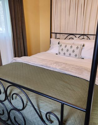 A neatly made bed with a green quilt, white blanket, decorative pillow, and black metal frame in a warmly lit bedroom with yellow walls and dark curtains. , The Goose Crown Inn