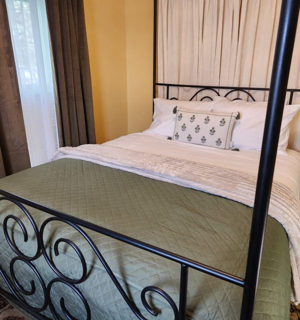 A neatly made bed with a green quilt, white blanket, decorative pillow, and black metal frame in a warmly lit bedroom with yellow walls and dark curtains. , The Goose Crown Inn