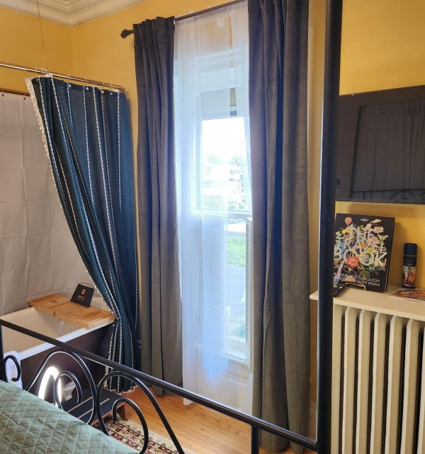 A bedroom with yellow walls, a four-poster bed, a clawfoot tub with a shower curtain, a window with dark curtains, and a radiator with books and a TV on top. , The Goose Crown Inn