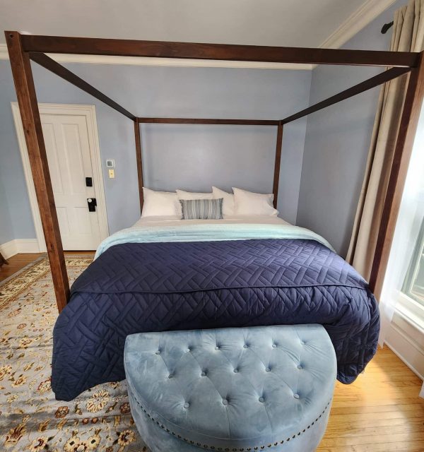 Four-poster bed with white linens and blue blanket, blue tufted bench at foot of bed, light blue walls, and a window with beige curtains. , The Goose Crown Inn