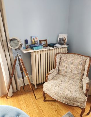 A vintage floral armchair sits on a hardwood floor next to a radiator topped with books, a framed photo, and decor; a tripod standing lamp is positioned nearby. , The Goose Crown Inn