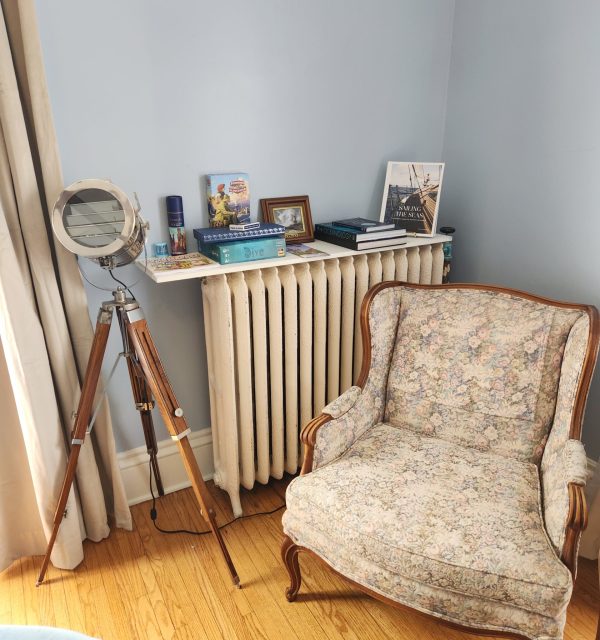 A vintage floral armchair sits on a hardwood floor next to a radiator topped with books, a framed photo, and decor; a tripod standing lamp is positioned nearby. , The Goose Crown Inn