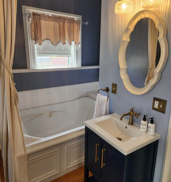 Bathroom with a white tub, a window with a pink valance, a navy vanity with a sink, gold fixtures, a mirror, and two glass light fixtures on a blue wall. , The Goose Crown Inn