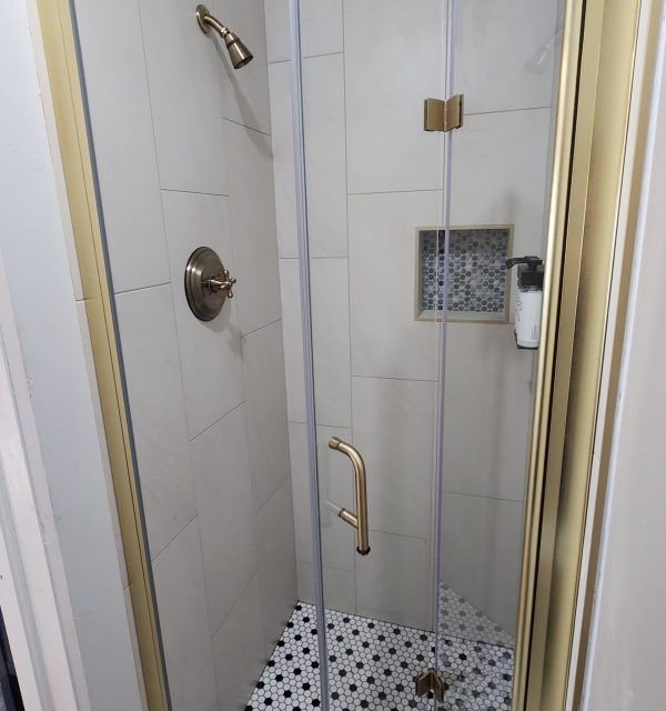 Small shower with beige tiled walls, gold hardware, glass door, built-in shelf with toiletries, and hexagonal black-and-white tile floor. , The Goose Crown Inn