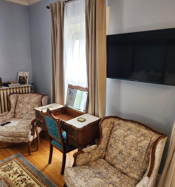 Living room with floral upholstered armchairs, a small wooden desk with a mirror, a wall-mounted TV, and light-colored curtains over a window. , The Goose Crown Inn