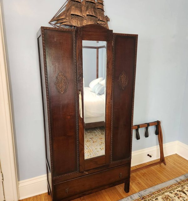 A dark wooden wardrobe with a mirrored door and carved detailing, topped with a wooden ship decoration, stands against a light blue wall on a hardwood floor. , The Goose Crown Inn