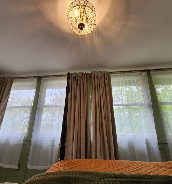 A bedroom with tall windows covered by sheer and heavy curtains, a ceiling light fixture, and part of a bed with green and orange bedding visible. , The Goose Crown Inn