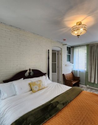 A bedroom with a four-poster bed, white brick wall, gold light fixture, curtained windows, an armchair, and a green blanket over an orange quilt. , The Goose Crown Inn