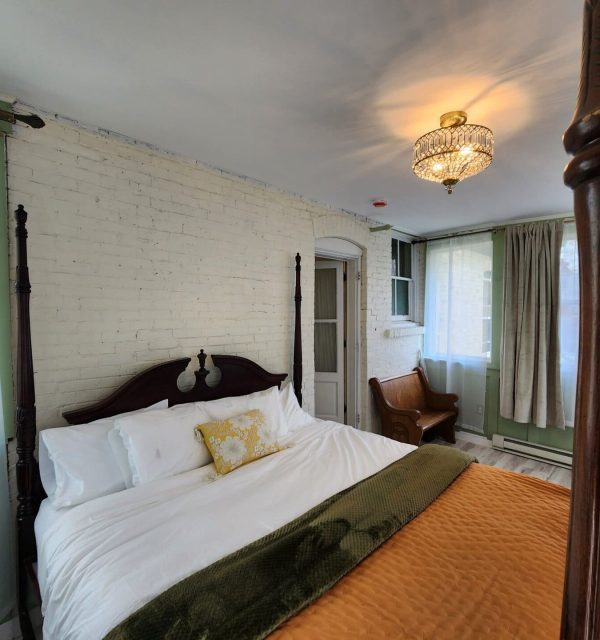 A bedroom with a four-poster bed, white brick wall, gold light fixture, curtained windows, an armchair, and a green blanket over an orange quilt. , The Goose Crown Inn