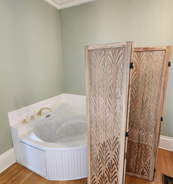A corner bathtub with brass fixtures is set against a light green wall, with a decorative wooden folding screen placed in front of it on a hardwood floor. , The Goose Crown Inn