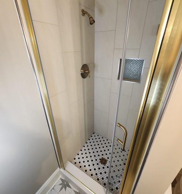 A small shower with gold trim, wall-mounted showerhead and handle, built-in niche, and black-and-white patterned tile floor. The bathroom floor has a starburst tile design. , The Goose Crown Inn