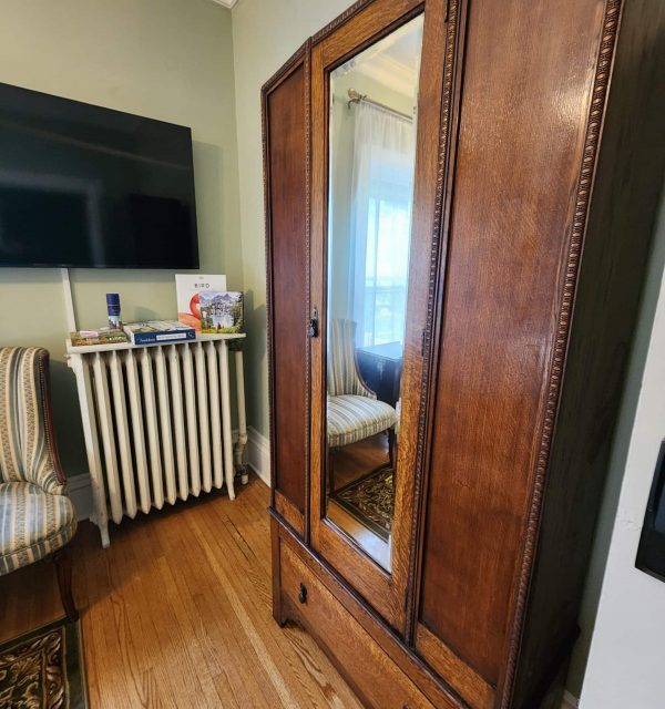 A wooden wardrobe with a mirrored door stands on a hardwood floor near a TV, radiator, and striped chairs in a room with green walls. , The Goose Crown Inn