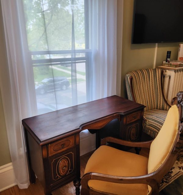 A small wooden desk with an ornate chair is placed in front of a window with sheer curtains; nearby are an upholstered chair, radiator, books, and part of a TV screen. , The Goose Crown Inn